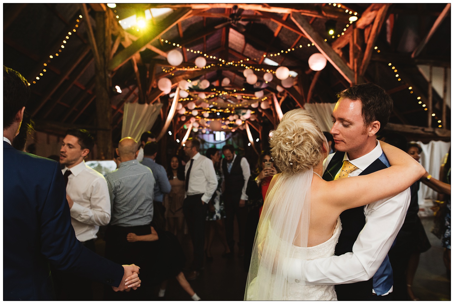 Fairly lights and paper lanterns decorate the barn in Alpheton at a wedding reception. The newly married couple dance in the foreground.