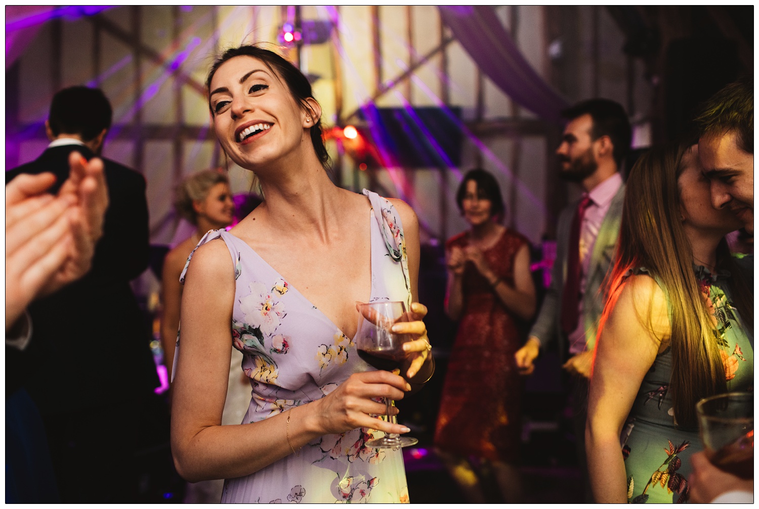 Woman with dark hair and a lila floral dress is holding a glass of red wine and smiling at a wedding reception.