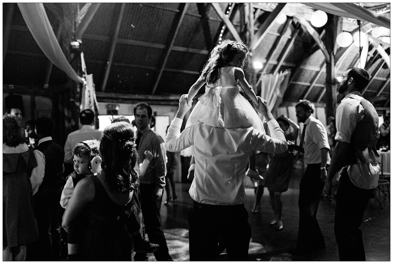 Small girl in a white dress on the shoulders of a man, a young boy wearing ear defenders, on the dancefloor at Alpheton.