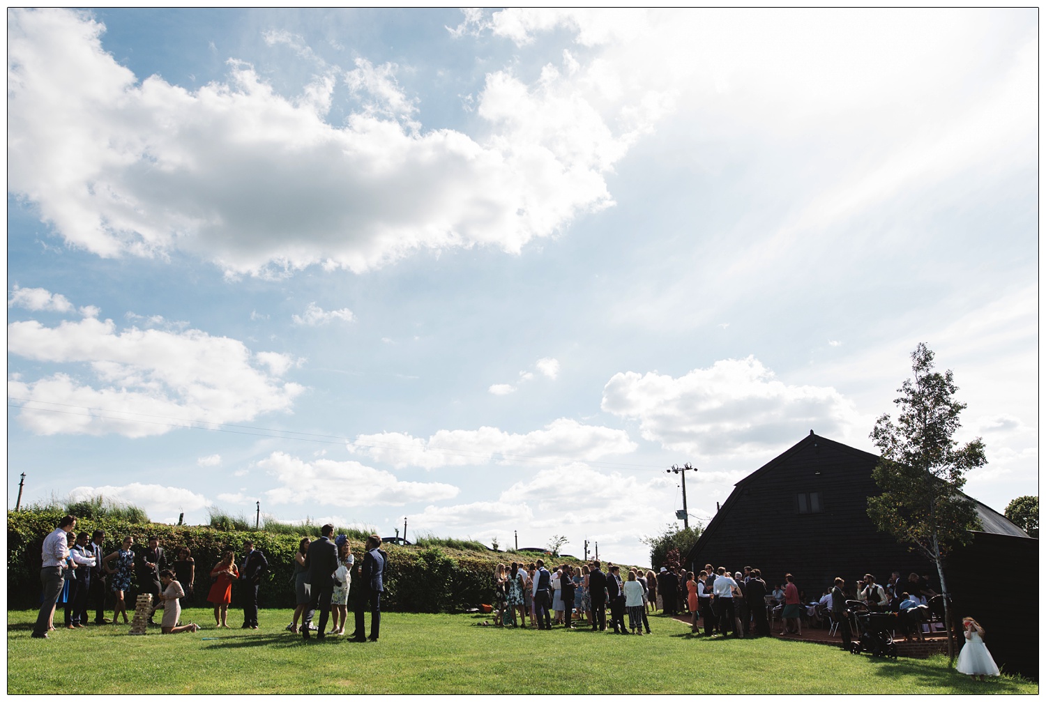 Guests in the stackyard wedding garden at Alpheton Hall Barns. The sky is blue with a few clouds. Guests are playing jenga and mingling, a small girl is taking photos.