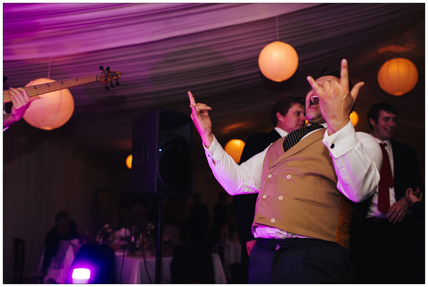 Man doing rock fingers,sign of the horns, in a marquee lit with purple lights at a wedding. He is wearing a waistcoat and tie.