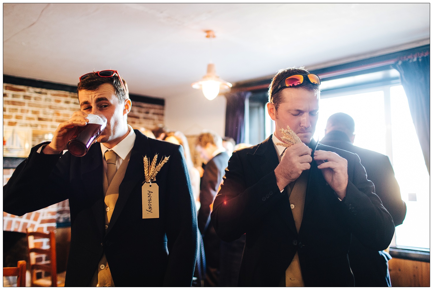 Groomsmen in the The Green Man Inn in Bradwell-on-Sea. One man is drinking a pint, the other is fixing his buttonhole, which is made of ears of wheat. They have sunglasses on their heads.