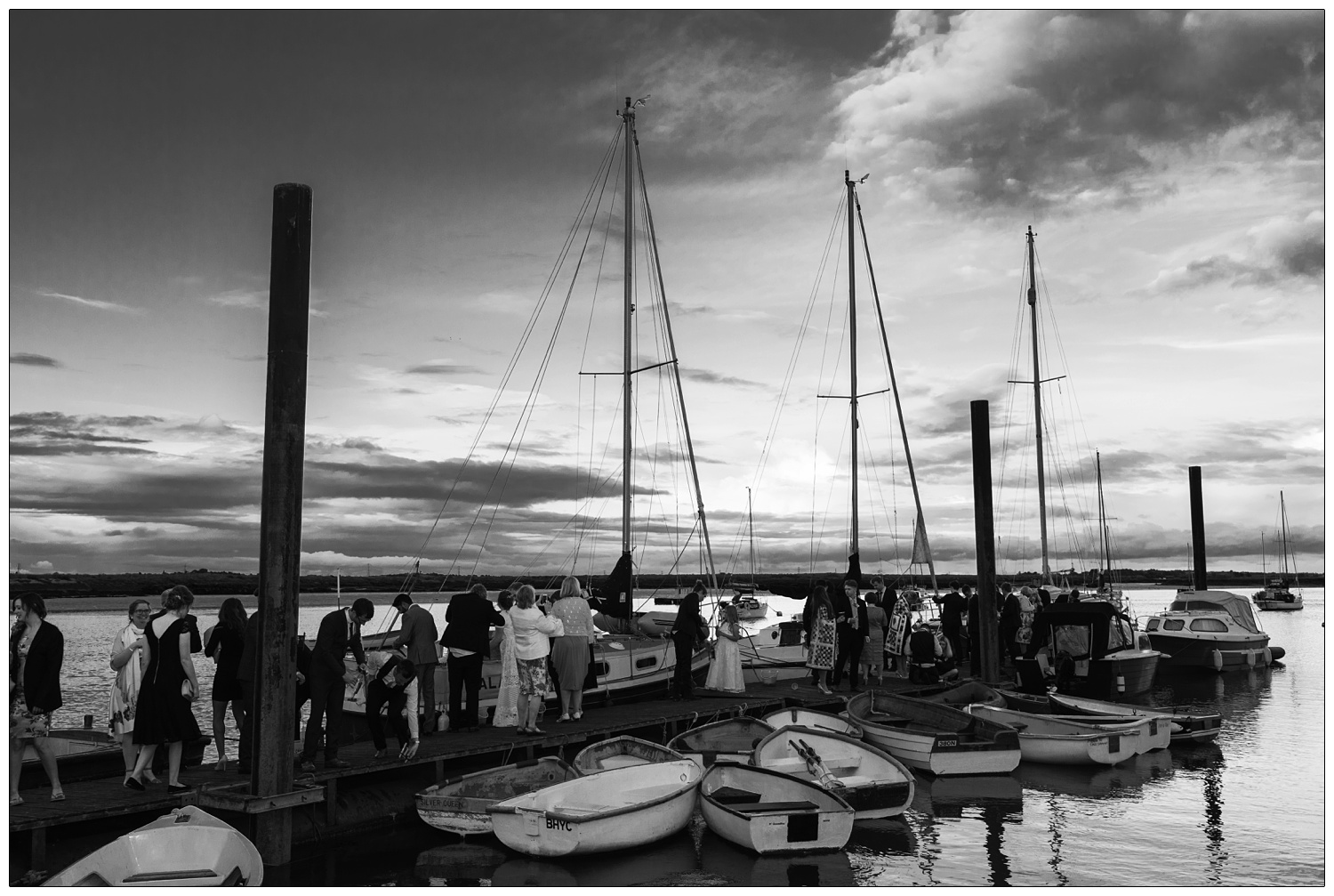 Wedding guests out on the jetty at the Brandy Hole venue. There are boats in the water.