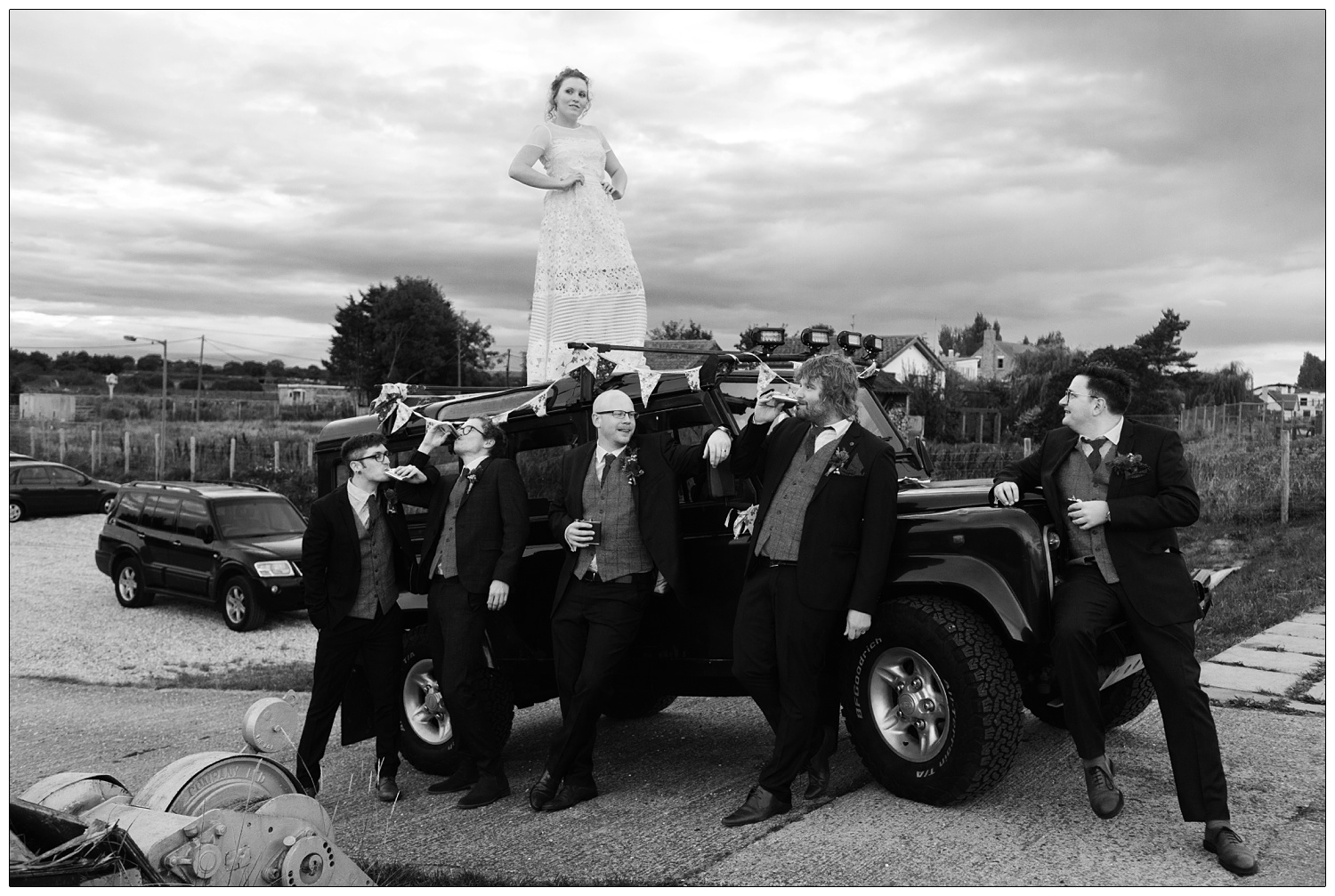 Bride with hands on hips stands on top of her Land Rover. Groom and groomsmen lean against it.