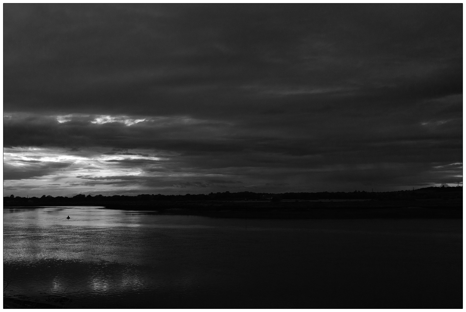A evening photograph taken from the Brandy Hole overlooking the River Crouch. South Woodham Ferrers is across the river. A gap of light in the clouds is reflected in the river. A black and white Essex landscape photograph.