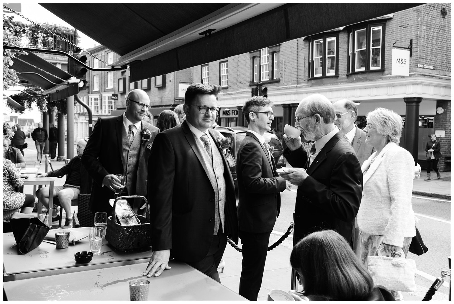 Bestman looking along the High Street with his hand on the table. The groom is behind him. One man is drinking tea. They are outside OAKhouse Bar Cafe in Maldon.