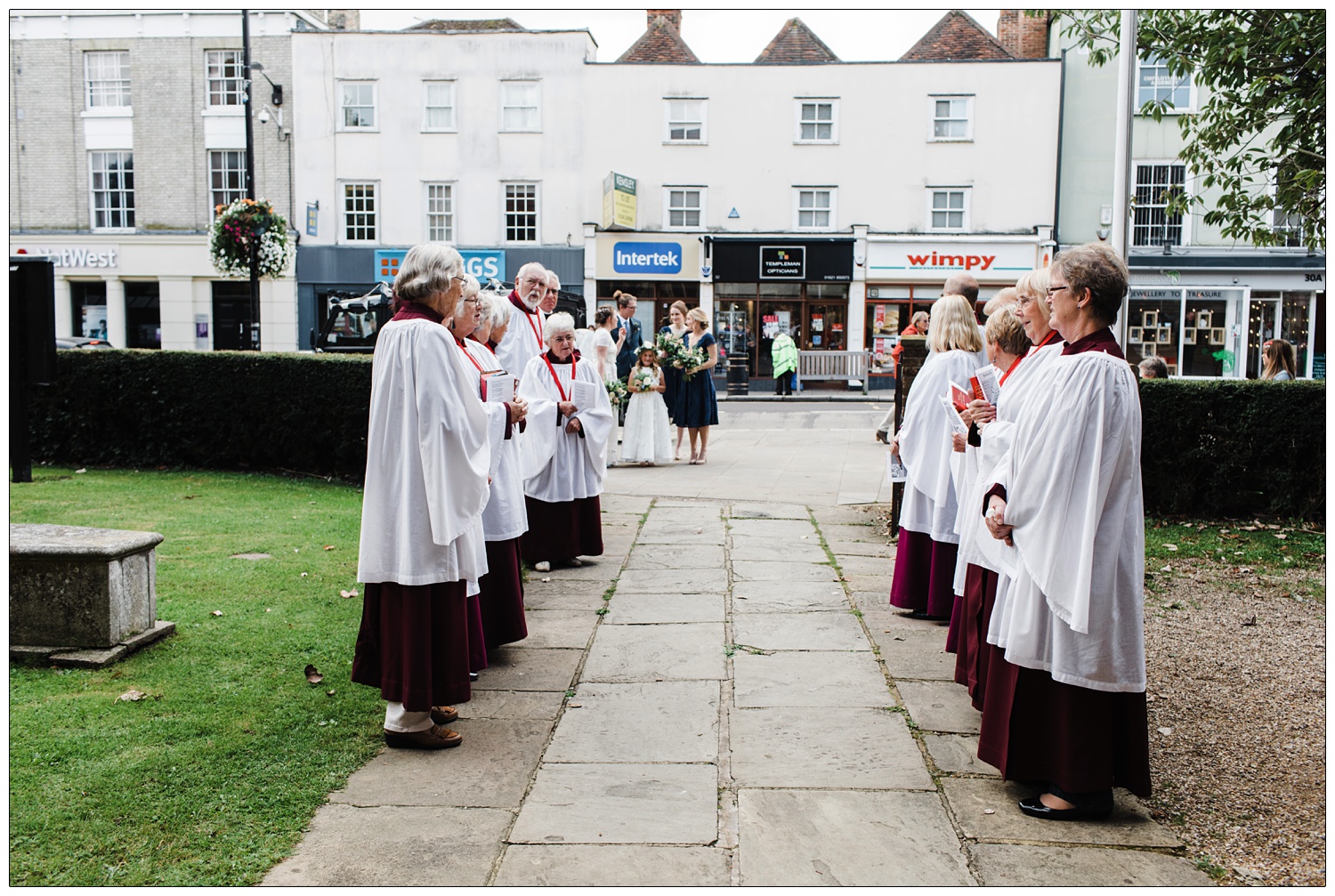 The choir line up outside All Saints with St Peter Church in Maldon. Waiting for the bride and bridesmaids to walk down the path. There is a Wimpy restaurant and opticians in the background.