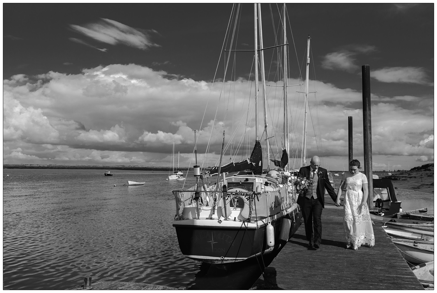 Bride and groom walk along the jetty next to a boat on the River Crouch.