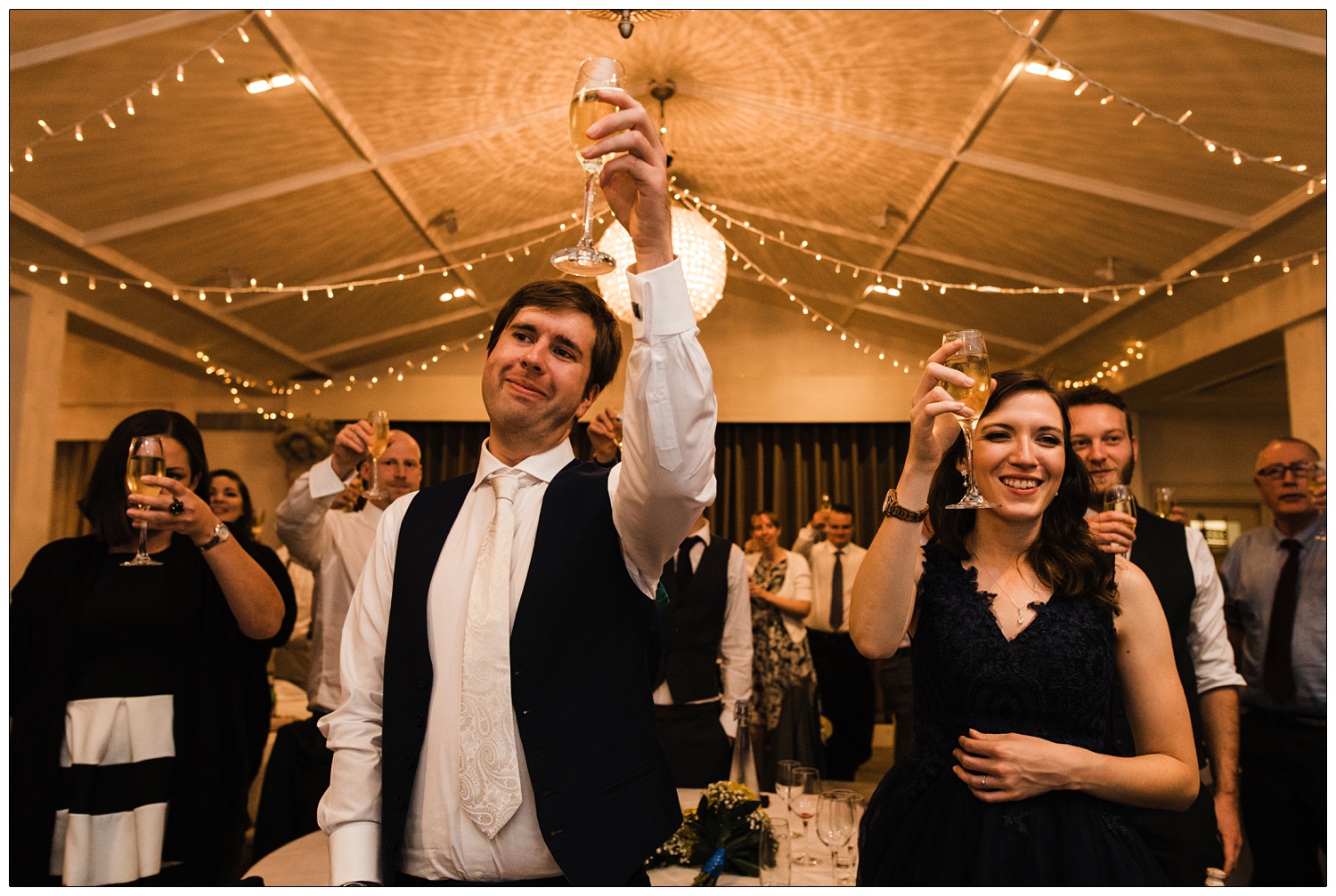 People stood around tables at a wedding reception in the ballroom. They are raising their glasses.