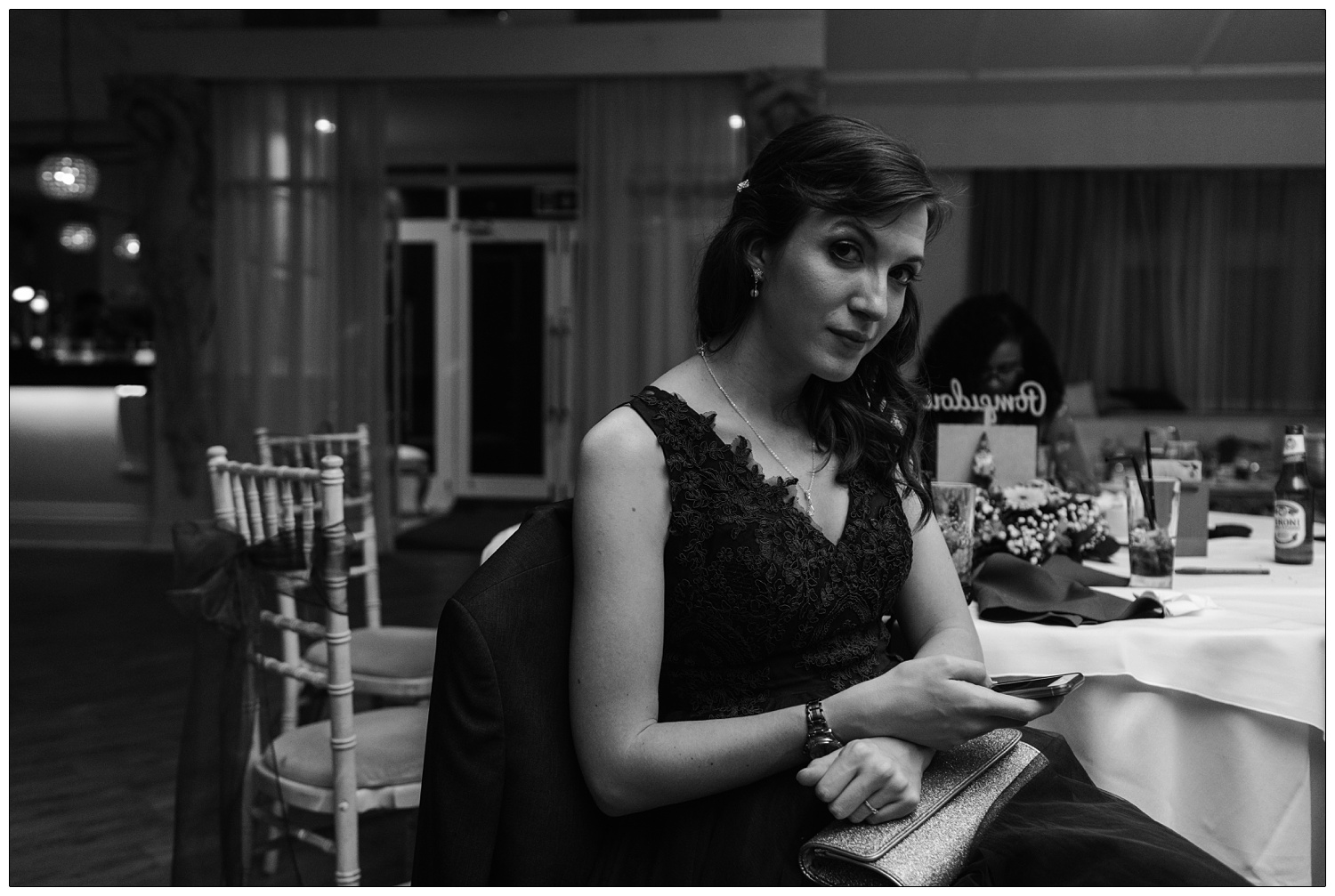 Bridesmaid at a wedding late at night, she's sat at a table checking her phone and looks up at the camera. A candid style wedding photograph.