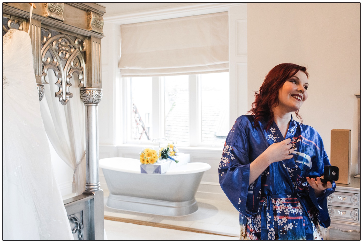 Bride in her blue floral dressing gown in a bedroom. The flowers are on top of the bath in the background. Her dress is hanging up on the four poster bed.