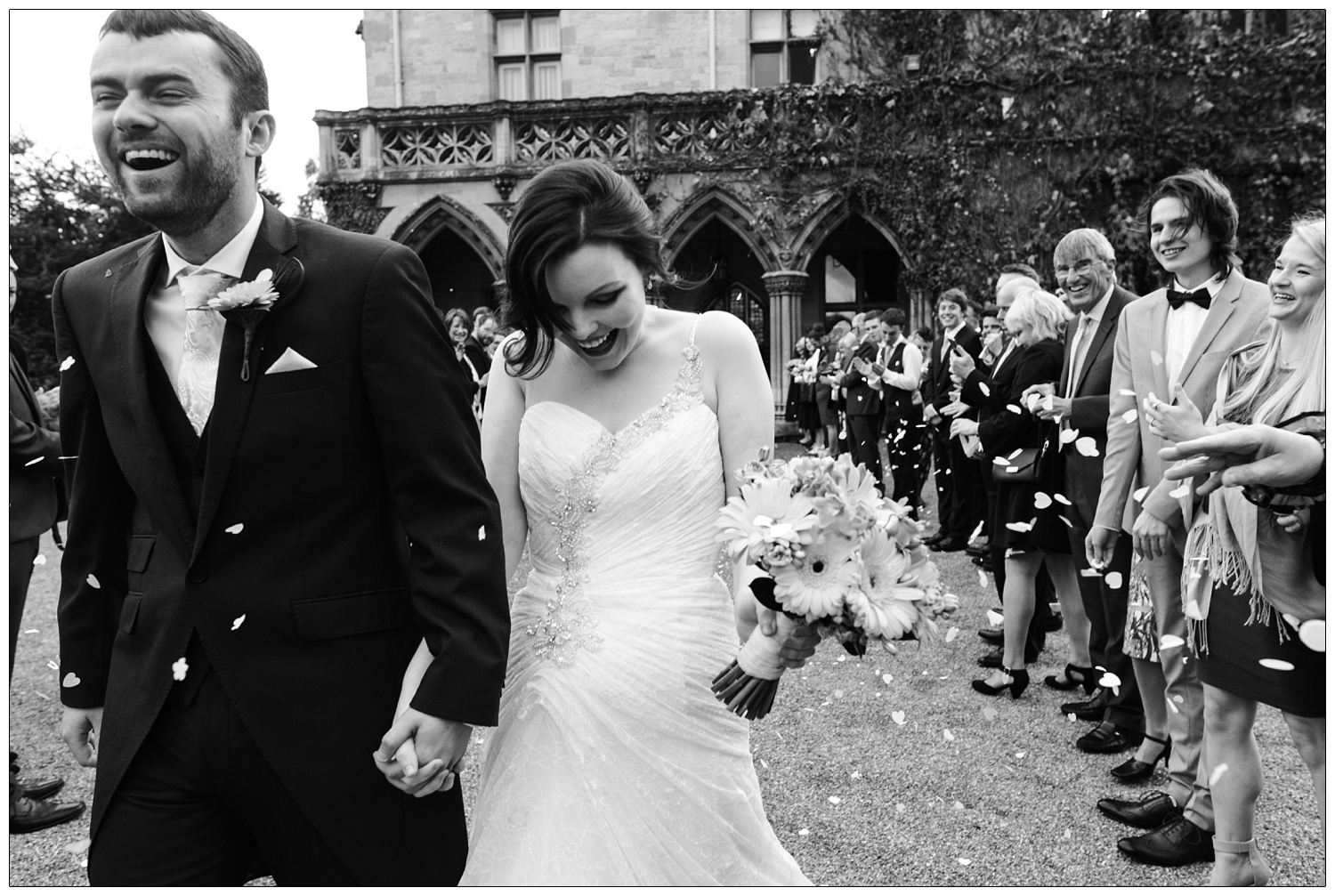 At Manor by the Lake wedding venue, a black and white photograph of man and woman holding hands. They just walked through a tunnel of guests throwing confetti.