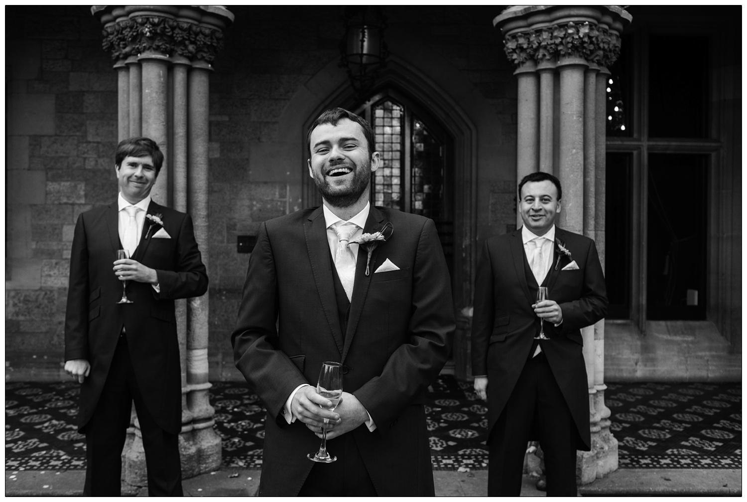 Black and white casual photo of groom with his groomsmen. He is laughing and they are all holding drinks.