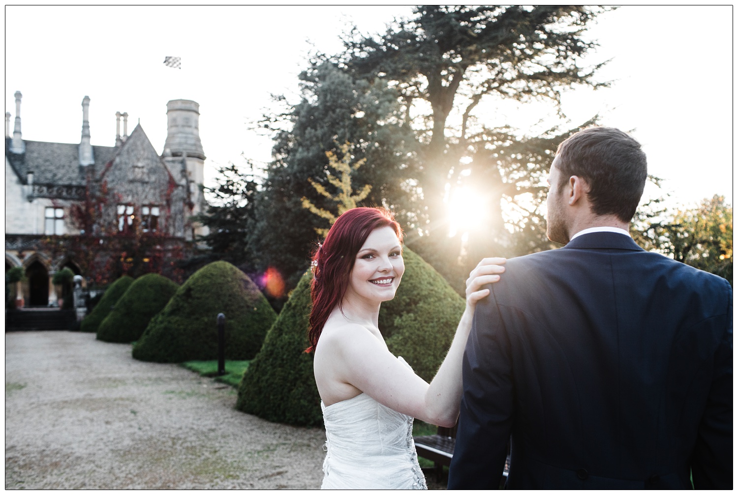Outside the Manor by the Lake venue, the October sunlight is soft and coming through the trees. Bride with red hair looks at the camera with her hand on her new husband's shoulder.
