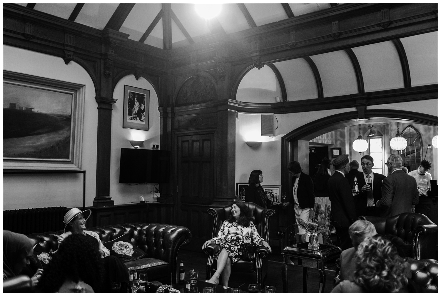 Wedding guests drinking and chatting. A woman is sat on a leather sofa in a hat, another woman is in a leather armchair and smiling. A natural reportage style wedding photograph at Manor by the Lake.