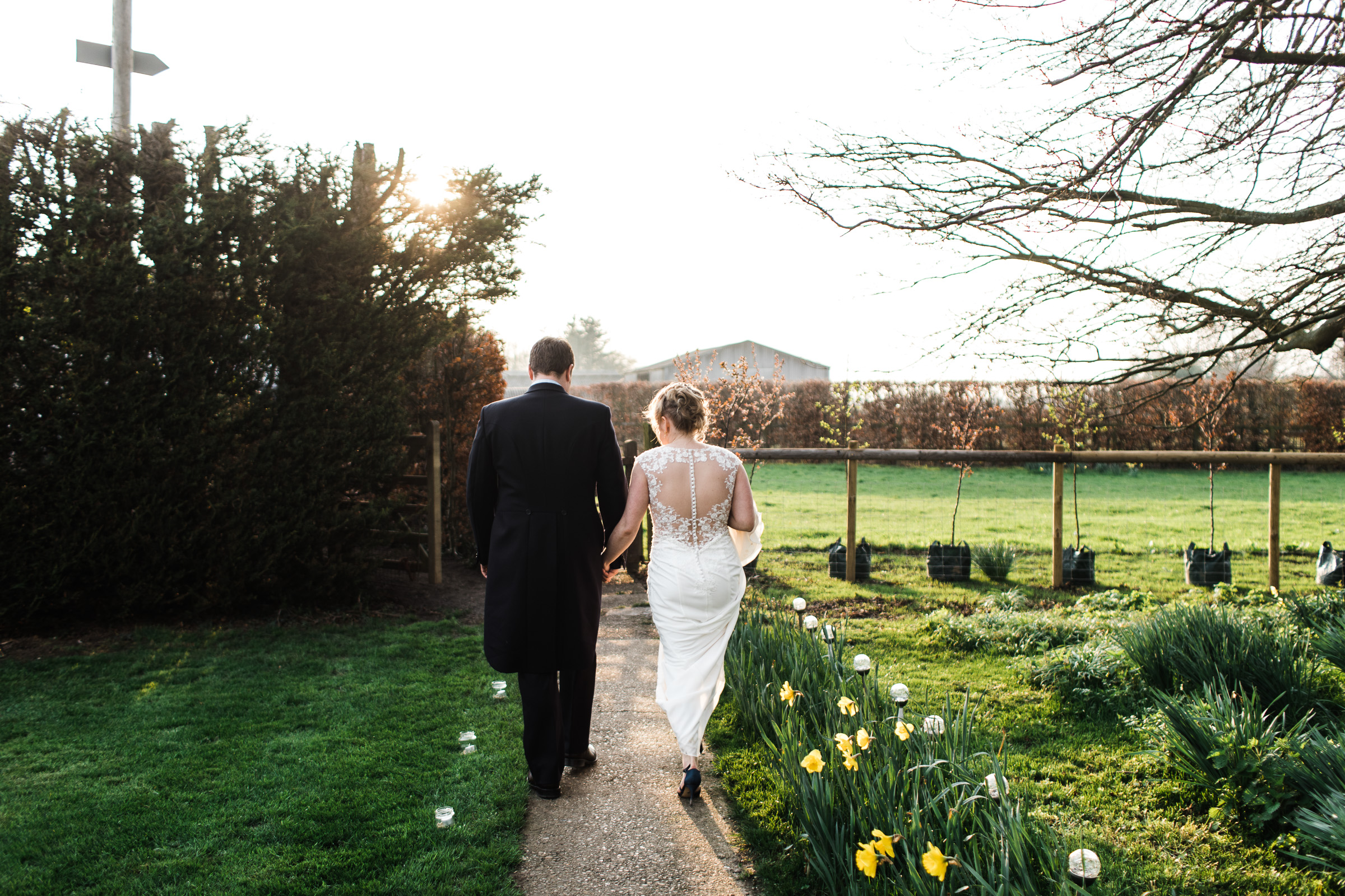 A spring farm wedding in Essex. The bride and groom walk down the path next to some daffodils in the gentle light from a sun low in the sky.