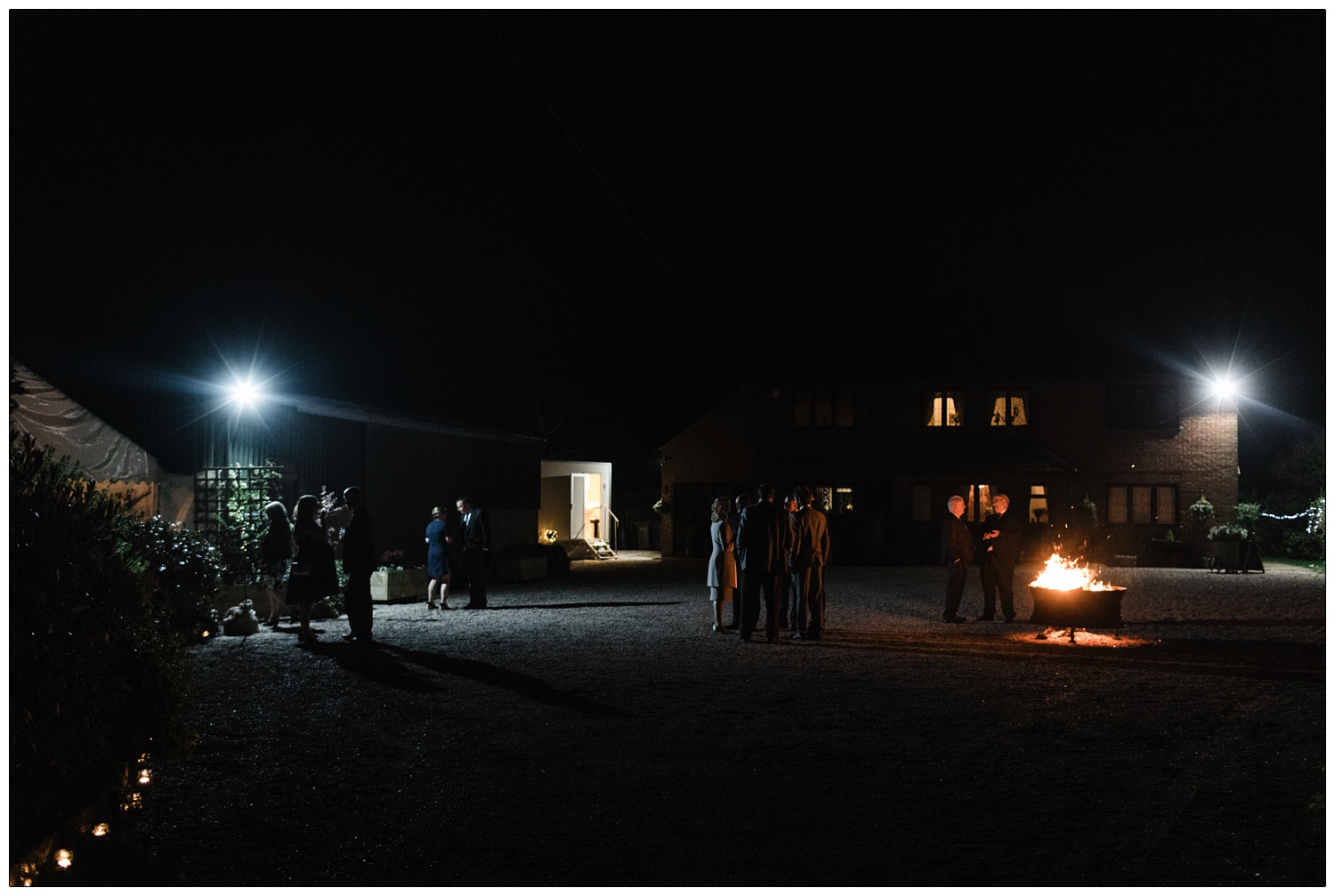 People outside at night gathered around a firepit in the garden outside the marquee and house.