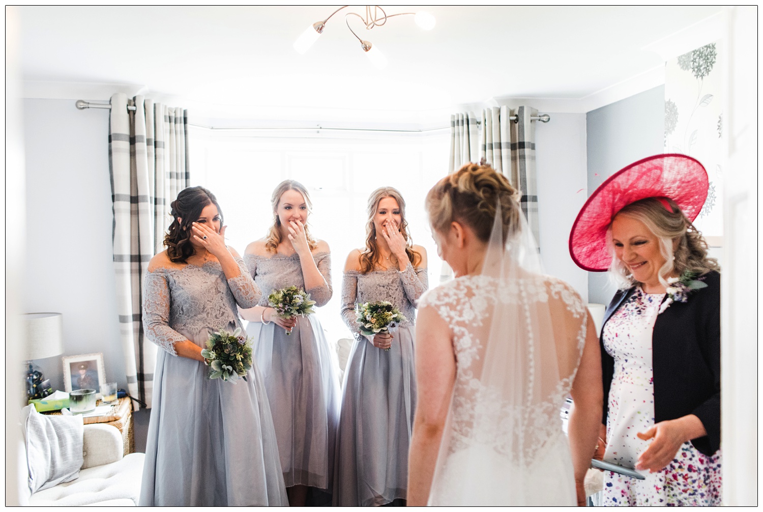 Bridesmaids overwhelmed as the bride reveals her dress in the living room. She's with her mum in a pink hat.