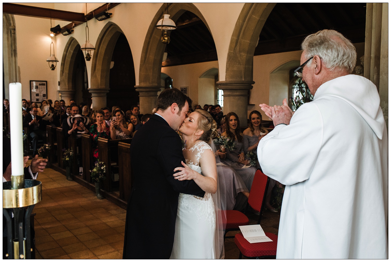 Bride and groom kissing now they are married. They are stood in front of the reverend who is clapping.