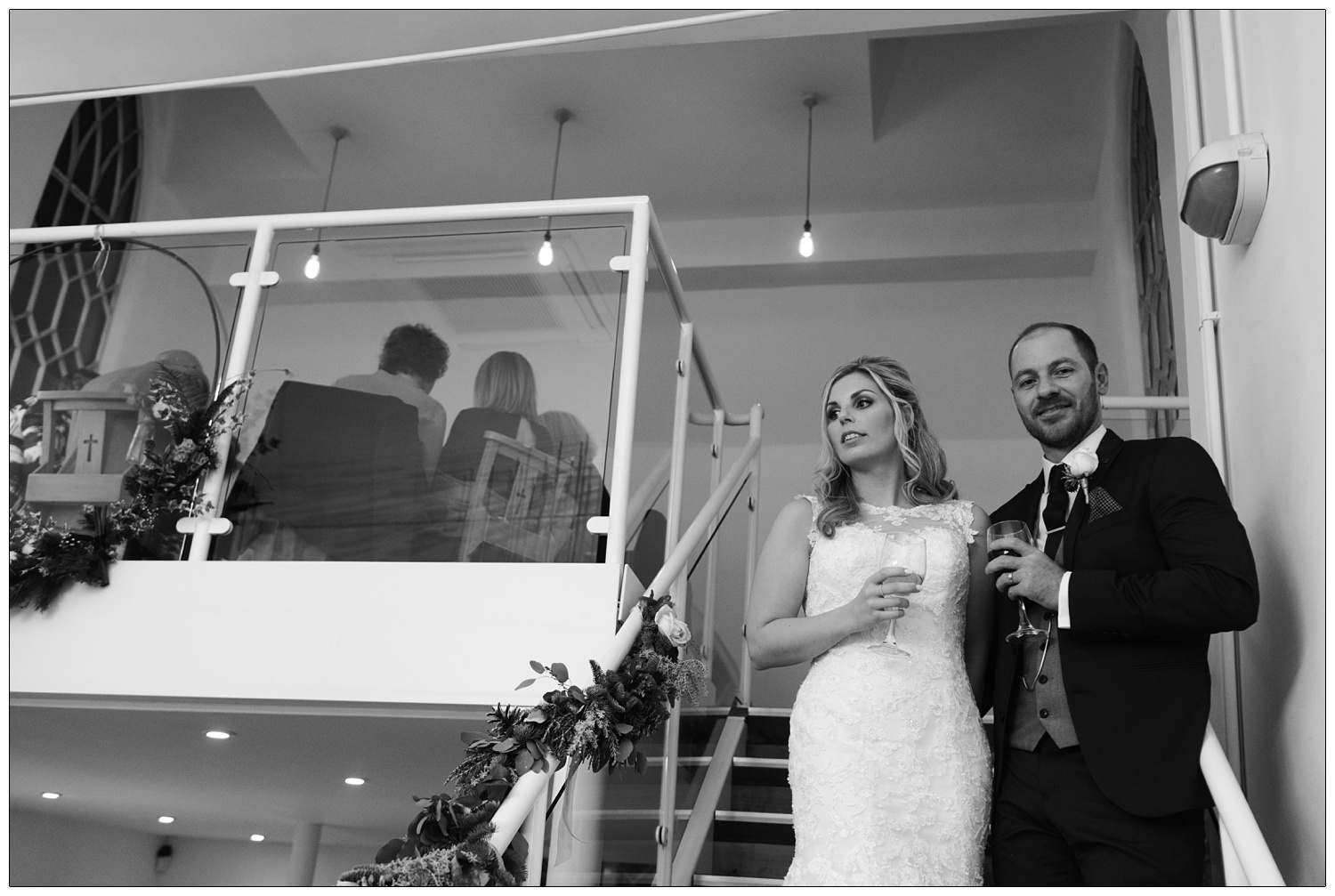 Bride and groom are on the stairs holding wine glasses. Groom is looking at the camera.