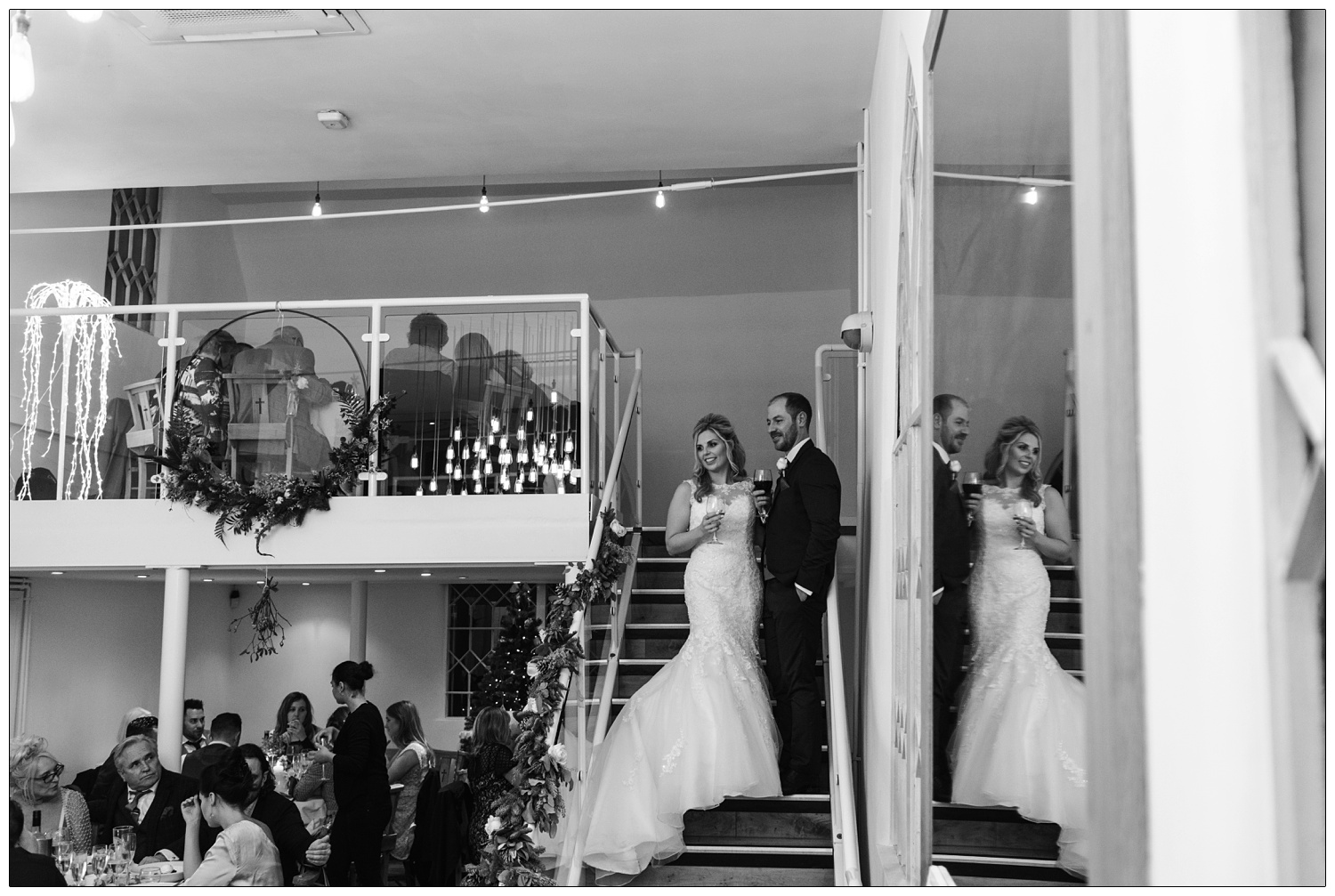 Bride and groom on the stairs of The Old Parish Rooms. They are reflected in a mirror. They are holding drinks and looking out over the tables of guests.