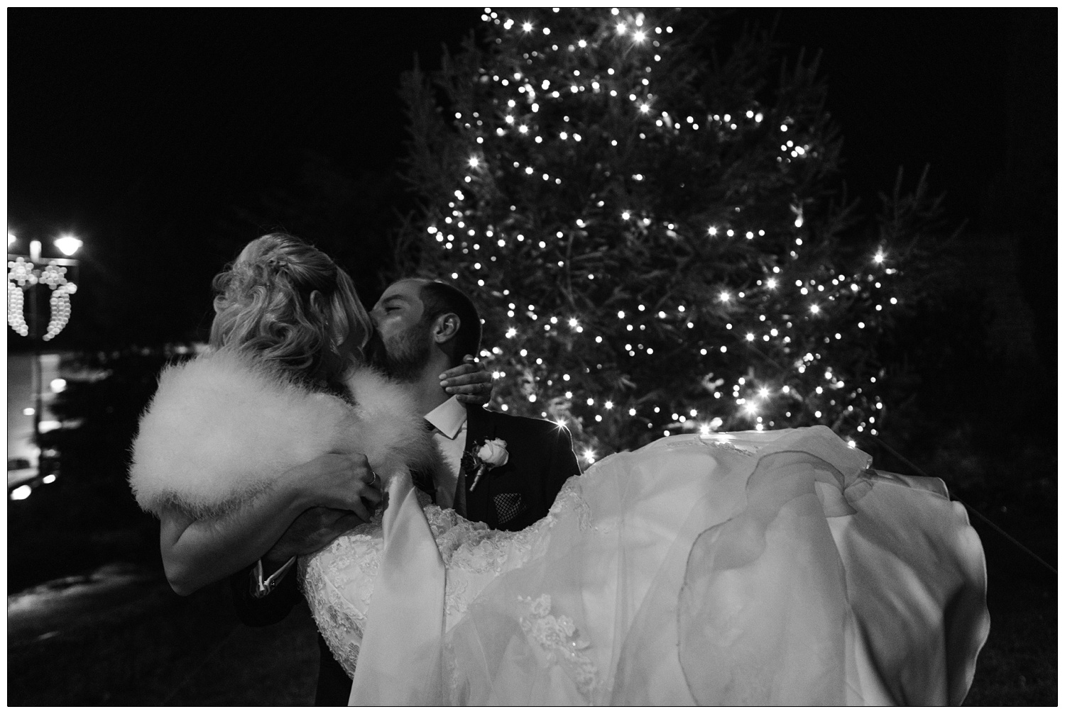 Man picks up woman in a wedding dress and kisses her outside and Christmas tree.