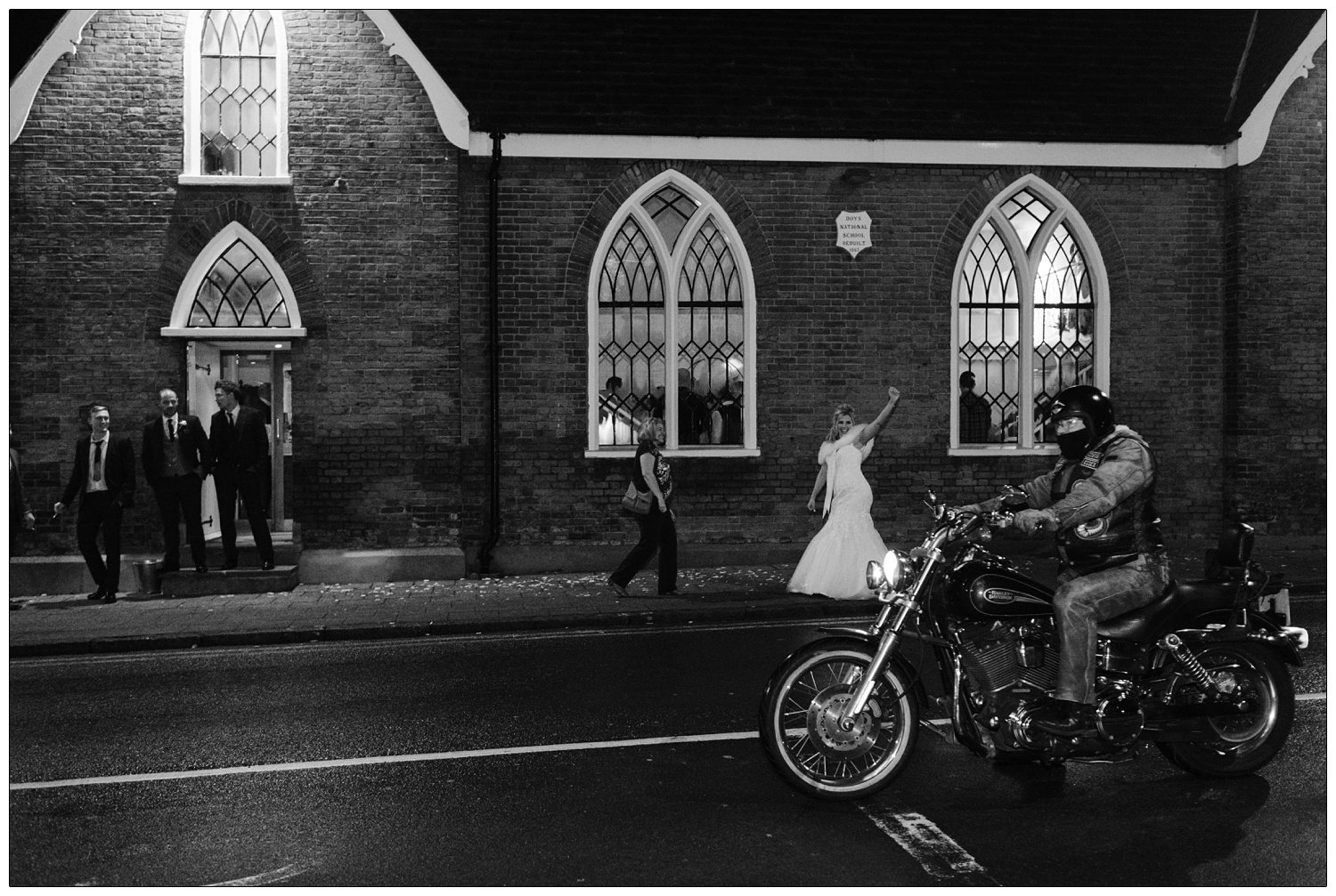 Bride raising her arm to greet the man on a motorbike riding past the Old Parish Rooms wedding venue in Rayleigh. The groom and two other men stand on the doorstep.