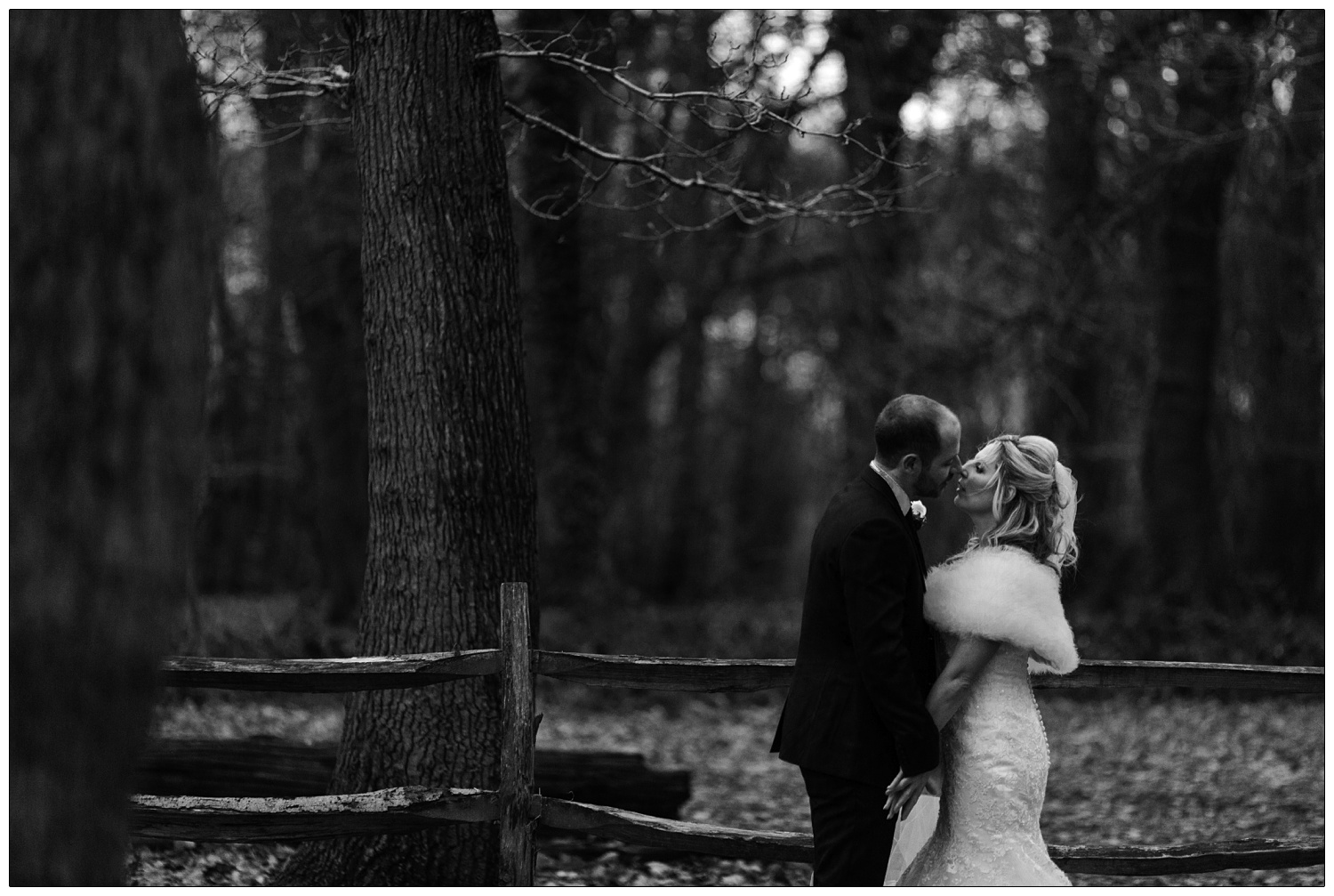 Newly married couple photographs in Hockley woods in December. They are facing each and are about to kiss. There are bare trees in the background and a small fence.