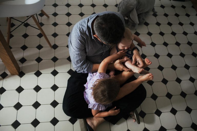 Man sat cross legged on a kitchen floor with his toddler and baby sat on his lap. Documentary family photography in London.