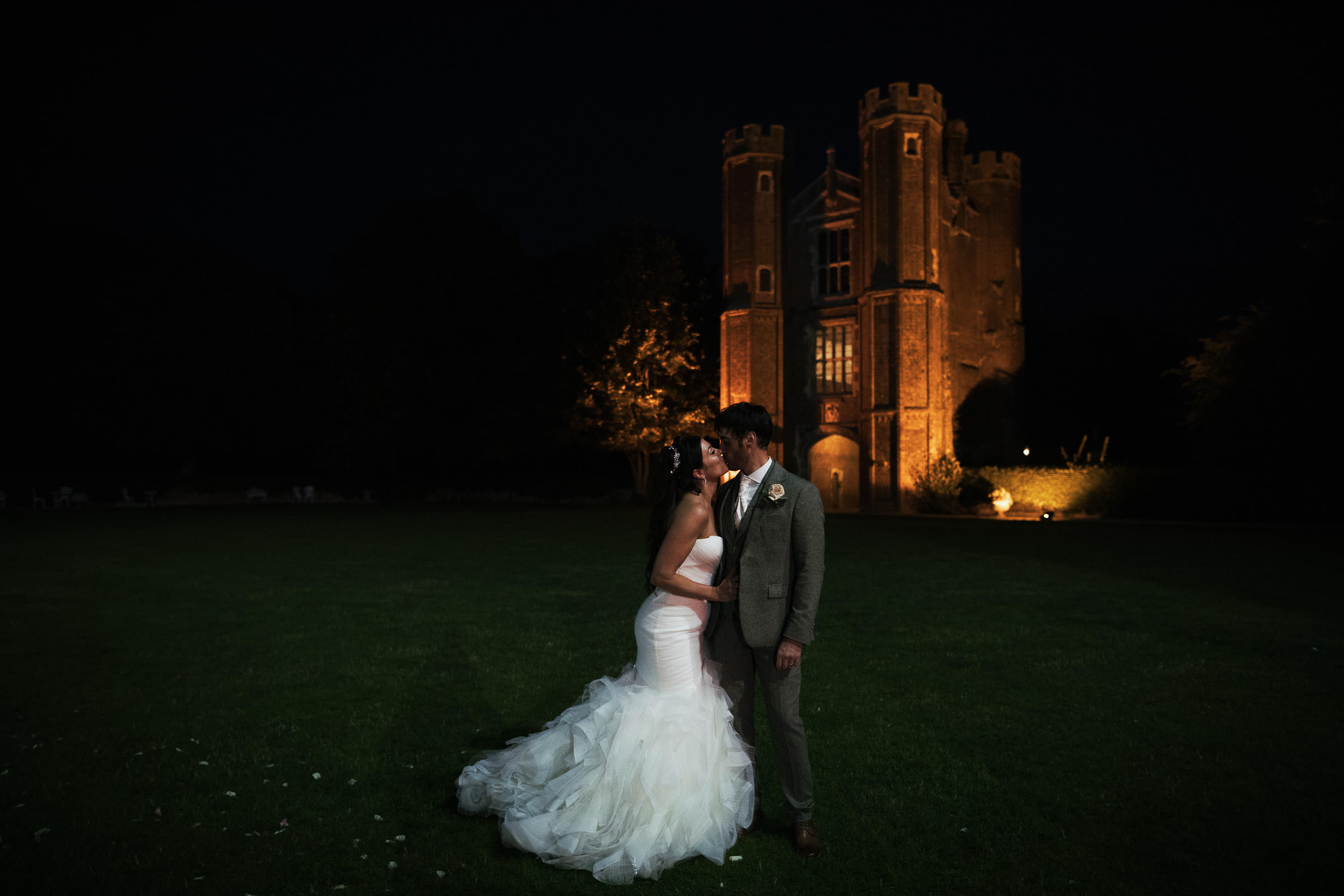 Newly married couple kiss on the lawn with the Great Tower at Leez Priory in the background. Lit up at night. Bride is wearing the Pronovias Barcelona Mildred Wedding Gown.
