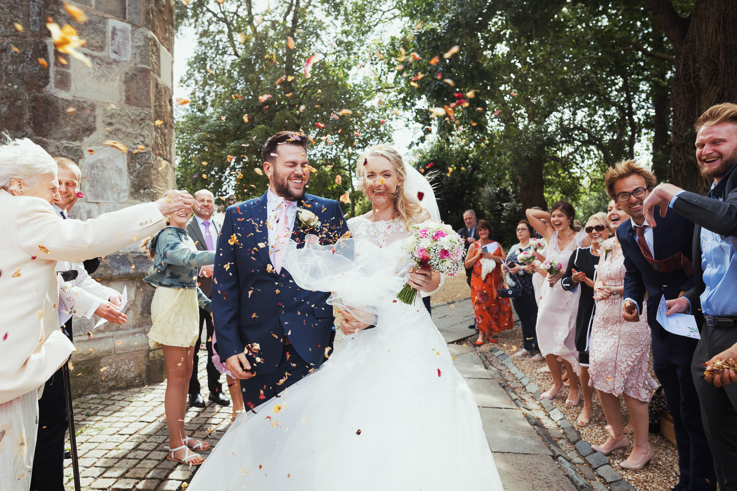 Wedding guests throw confetti petals at a couple after their wedding ceremony. It's a windy day.
