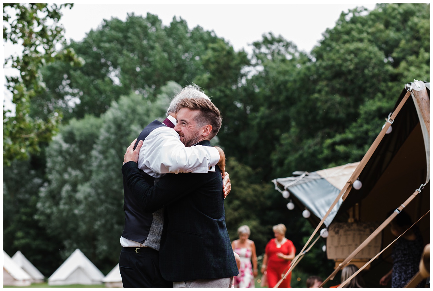 Two men hugging at a wedding. They are outside, there are trees behind them.