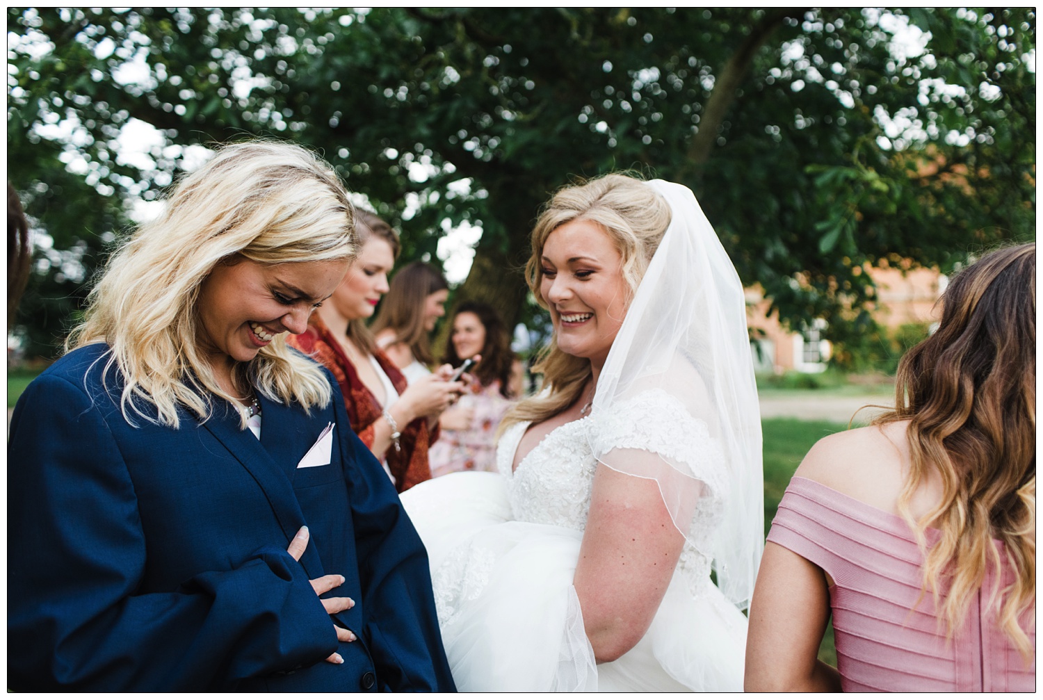 Bride is laughing with her friend. Her friend has put on a man's dark blue jacket to keep warm.