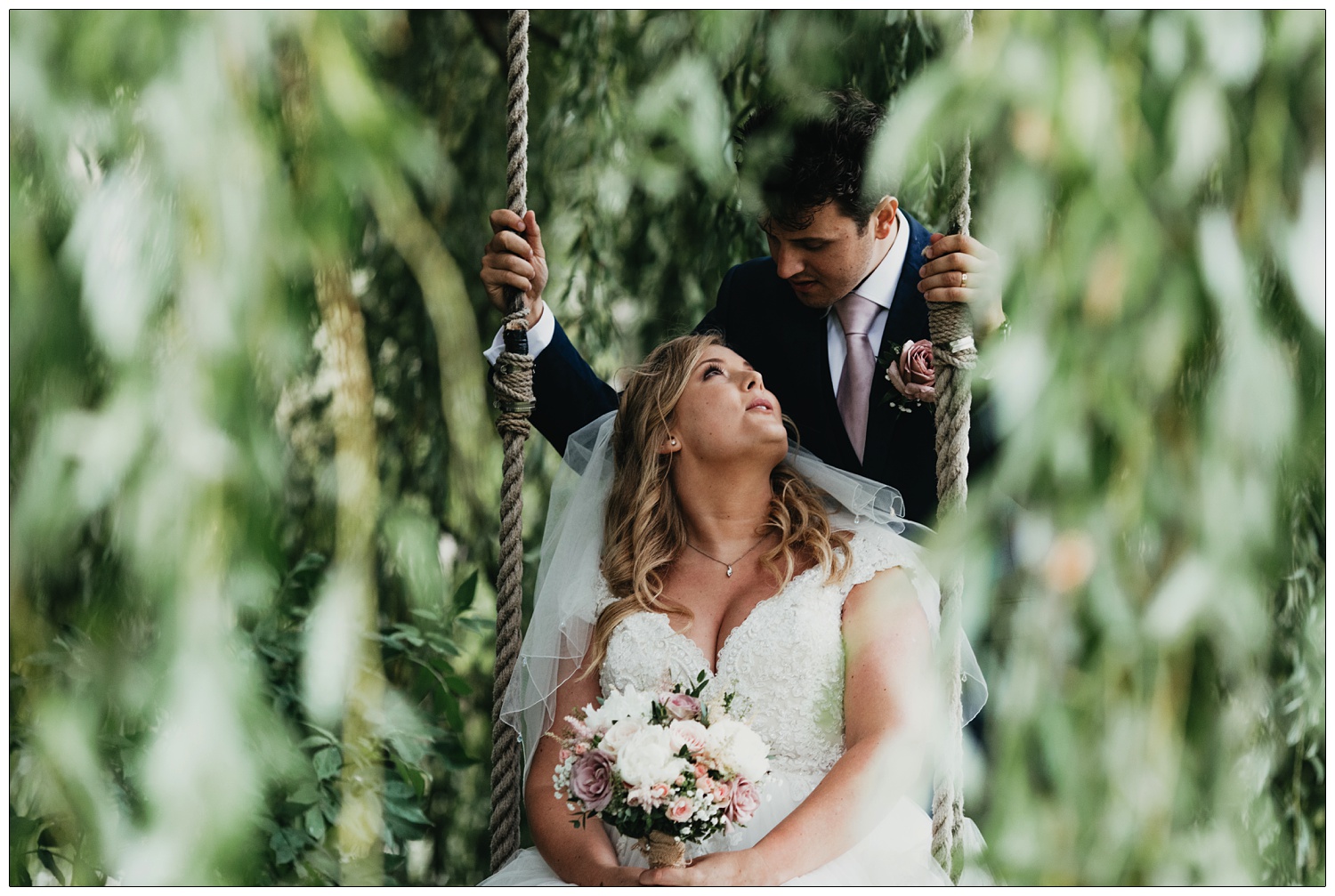 A bride sits on a swing hanging from a willow tree and her groom is behind her. She's looking up at him and holding her flowers. They are in the grounds of Anne of Cleves Barn Great Lodge wedding venue near Braintree.