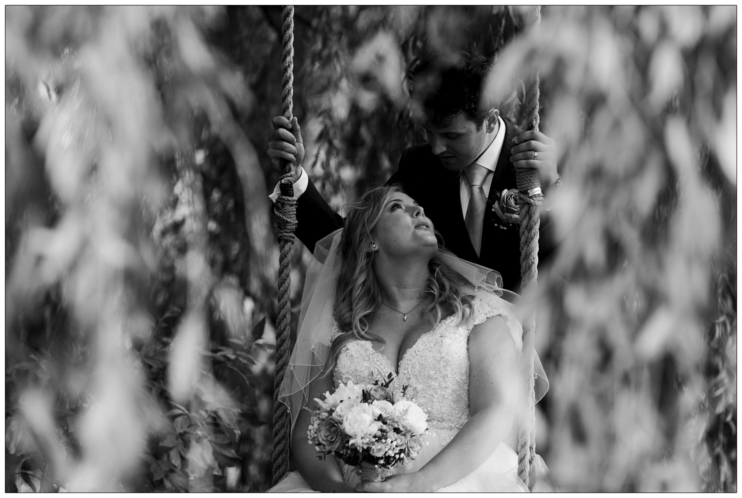 A bride sits on a swing hanging from a willow tree and her groom is behind her. She's looking up at him and holding her flowers. They are in the grounds of Anne of Cleves Barn Great Lodge wedding venue near Braintree.