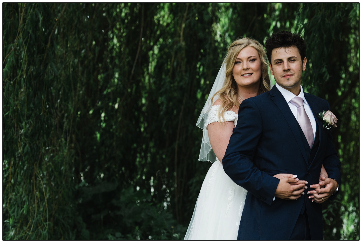 In front of a willow tree a man is standing in front of the woman he just married. She puts her arms around him. She has a white dress, veil and blonde hair. He is wearing a navy suit with a pale pinkish tie.