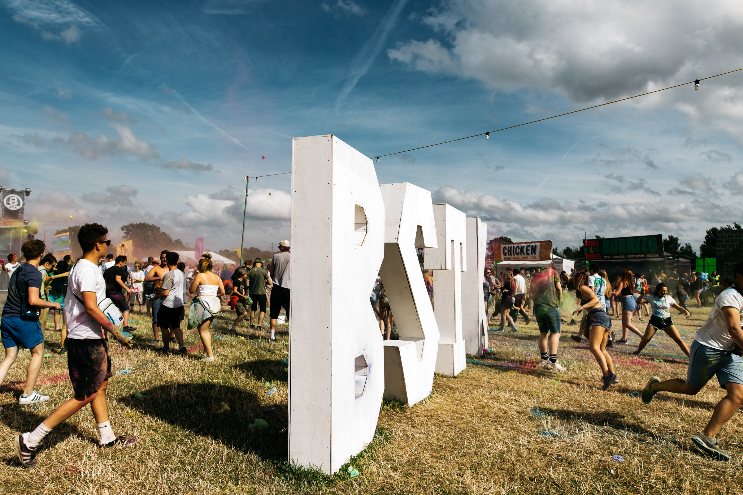 Giant BSTK letters in a field at a music festival. People are having a paint fight around it. Food stalls in the distance.