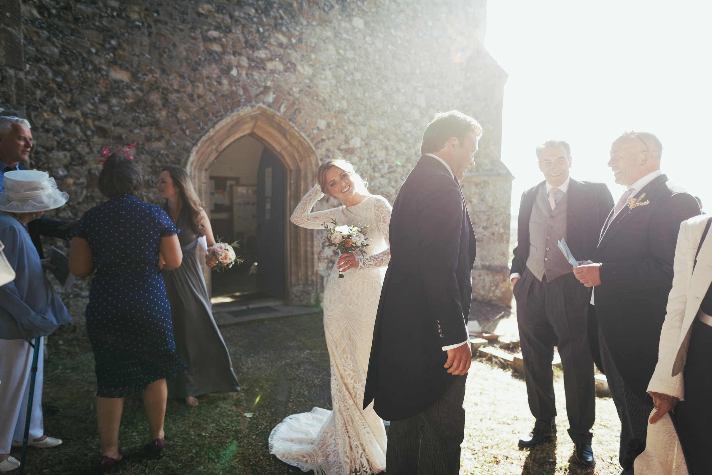 Wedding guests are chatting to each other but the bride is looking at the camera, illuminated by the sunlight. The door of The Church of St Mary & St Margaret in Stow Maries is behind her. She is wearing a long lace wedding dress with long sleeves.