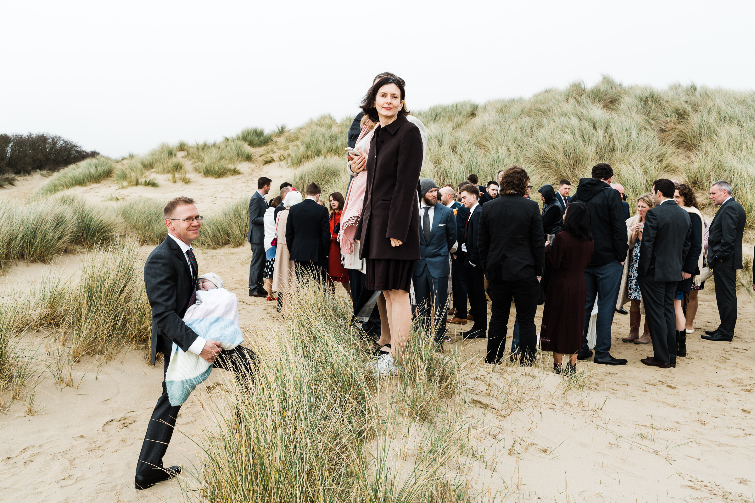 A woman standing on a sand dune and a man holding a baby. Wedding guests are in the background. They are on the beach at Camber Sands.