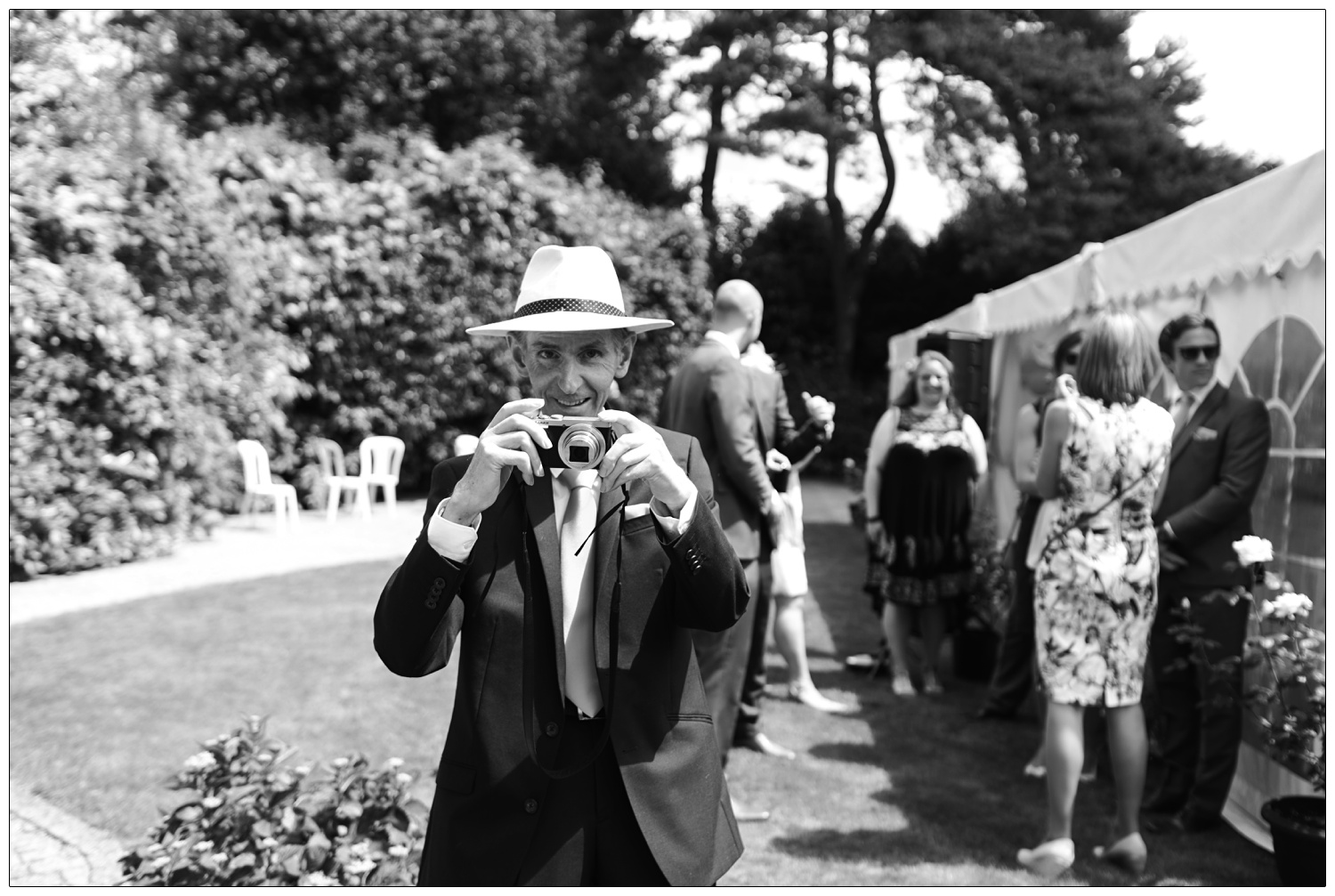 A man in a suit and hat in the sunshine, takes a photograph of the photographer.