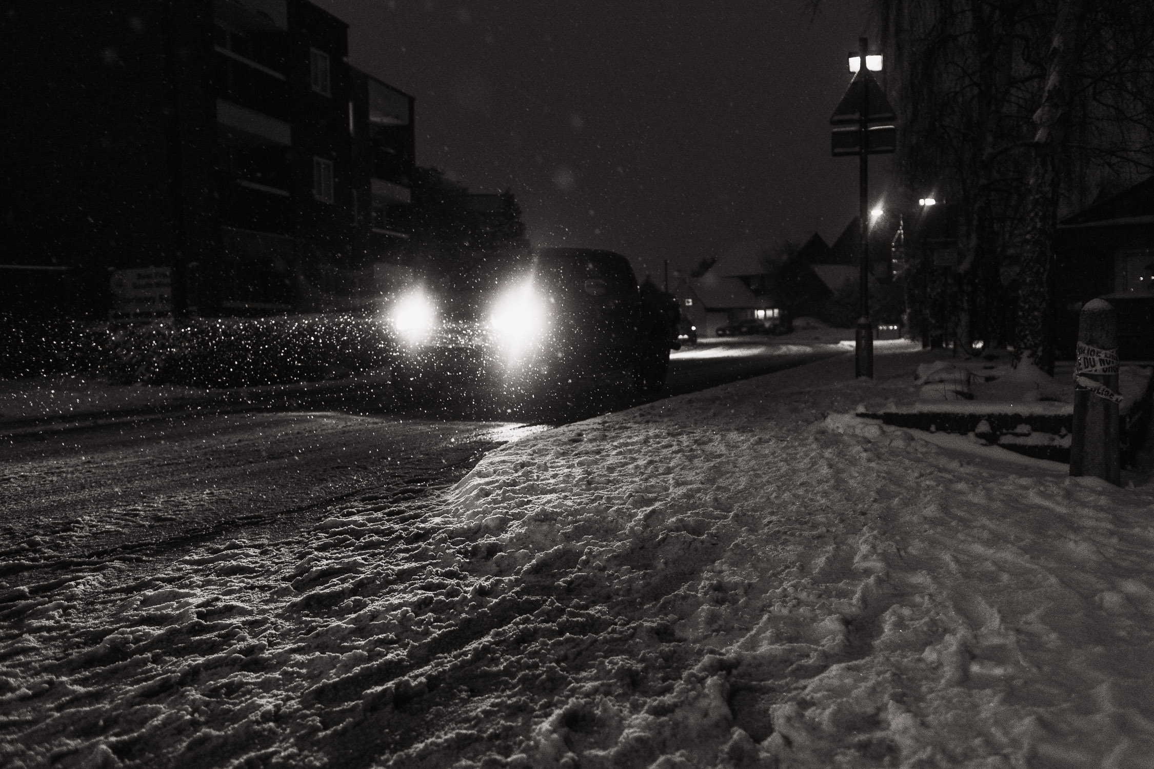 A car approaching the South Woodham Ferrers station on Hullbridge Road at night, amidst a snowy scene. In the background, The Cedars flats are visible.