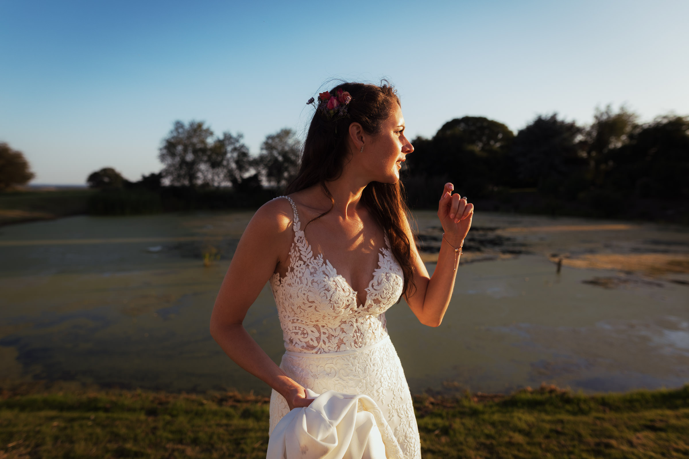 The bride in a lace wedding dress stood by a pond. The sun is setting so the light is low and casting shadows. The wedding is in Essex near South Woodham Ferrers. Candid wedding photographer from Essex. Bride is wearing Stella York 6648 from The Wedding Shop in Colchester.