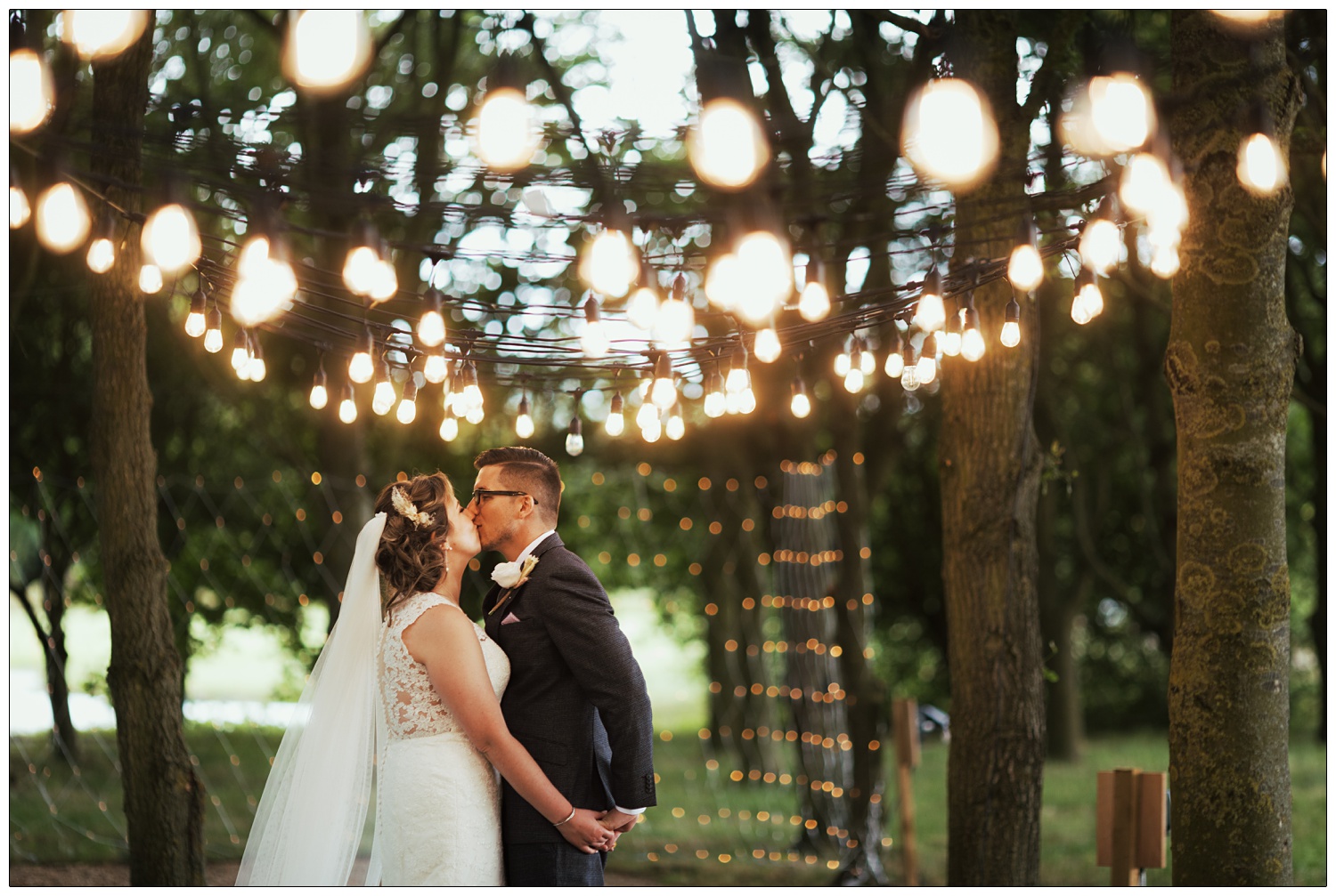 A newly married couple kiss among the trees under fairy lights. At Fynn Valley Terrace.
