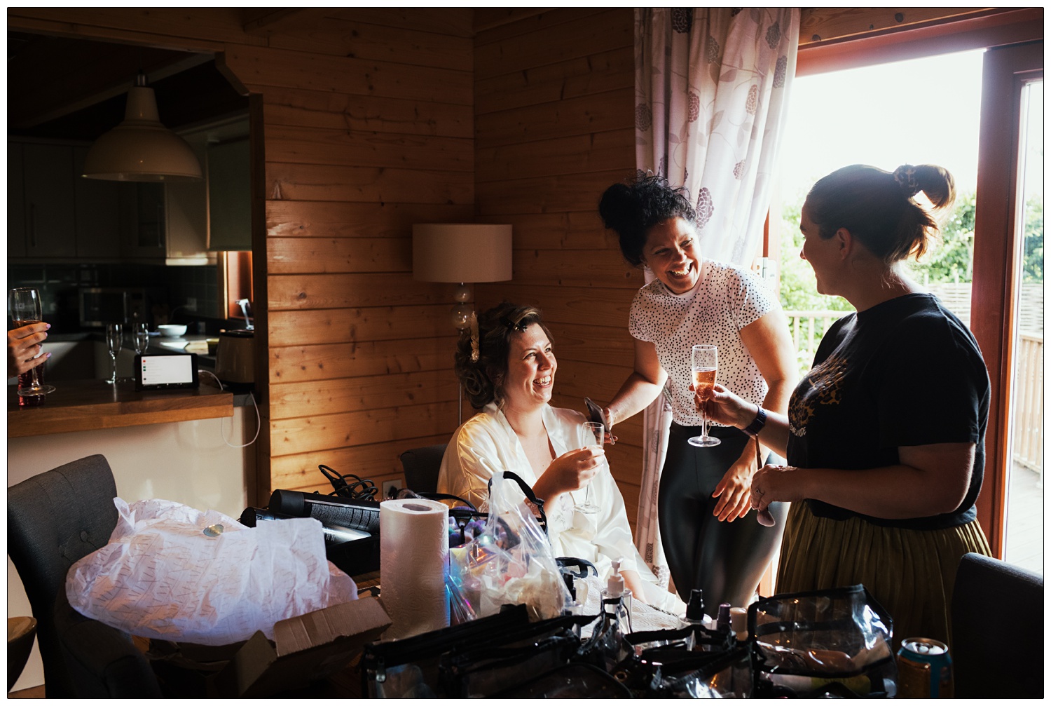 A woman in a white dressing gown raises a glass with a hair dresser and make up artist. They are laughing.