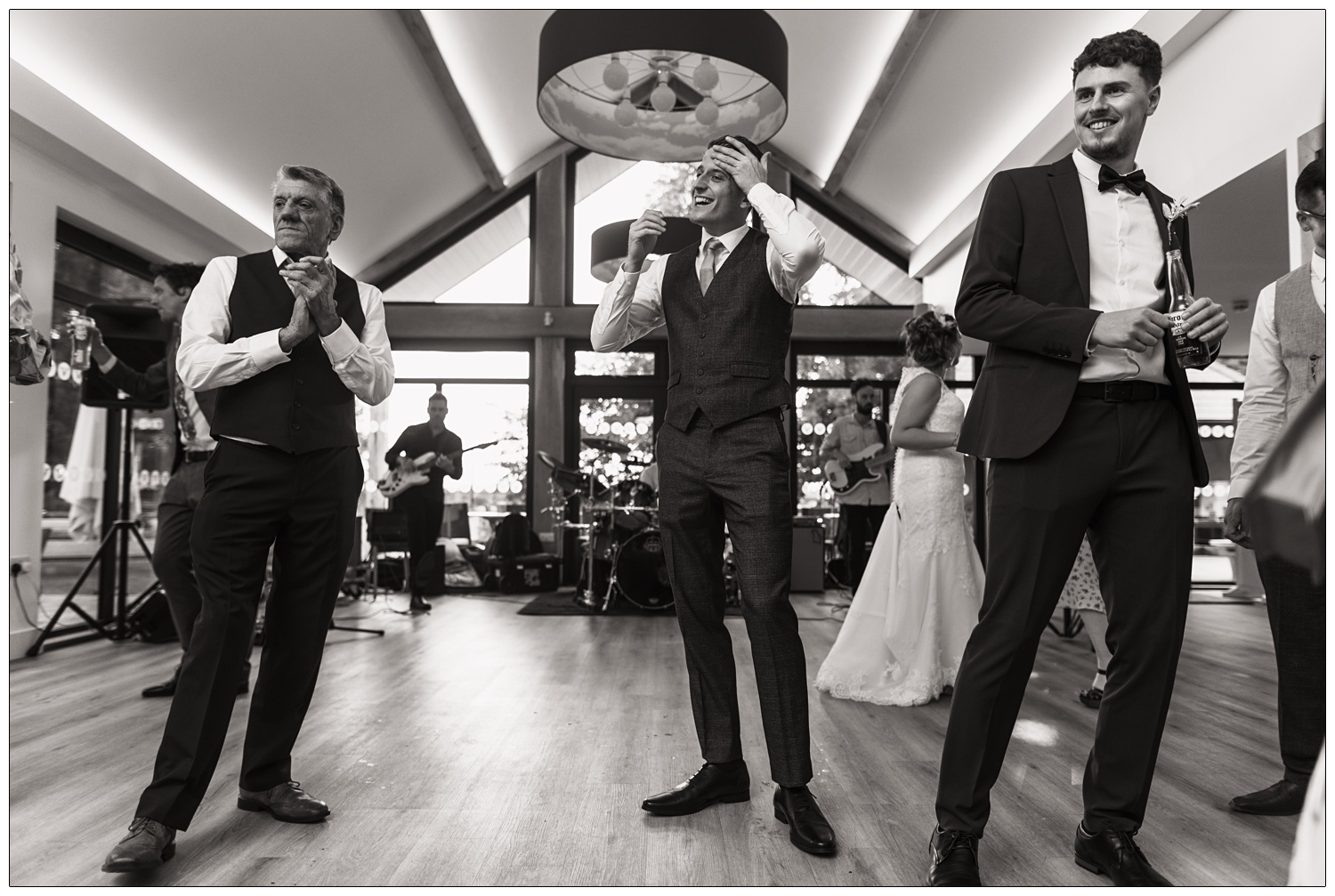 Three men in suits standing on a dance floor at a wedding. The band are behind them.