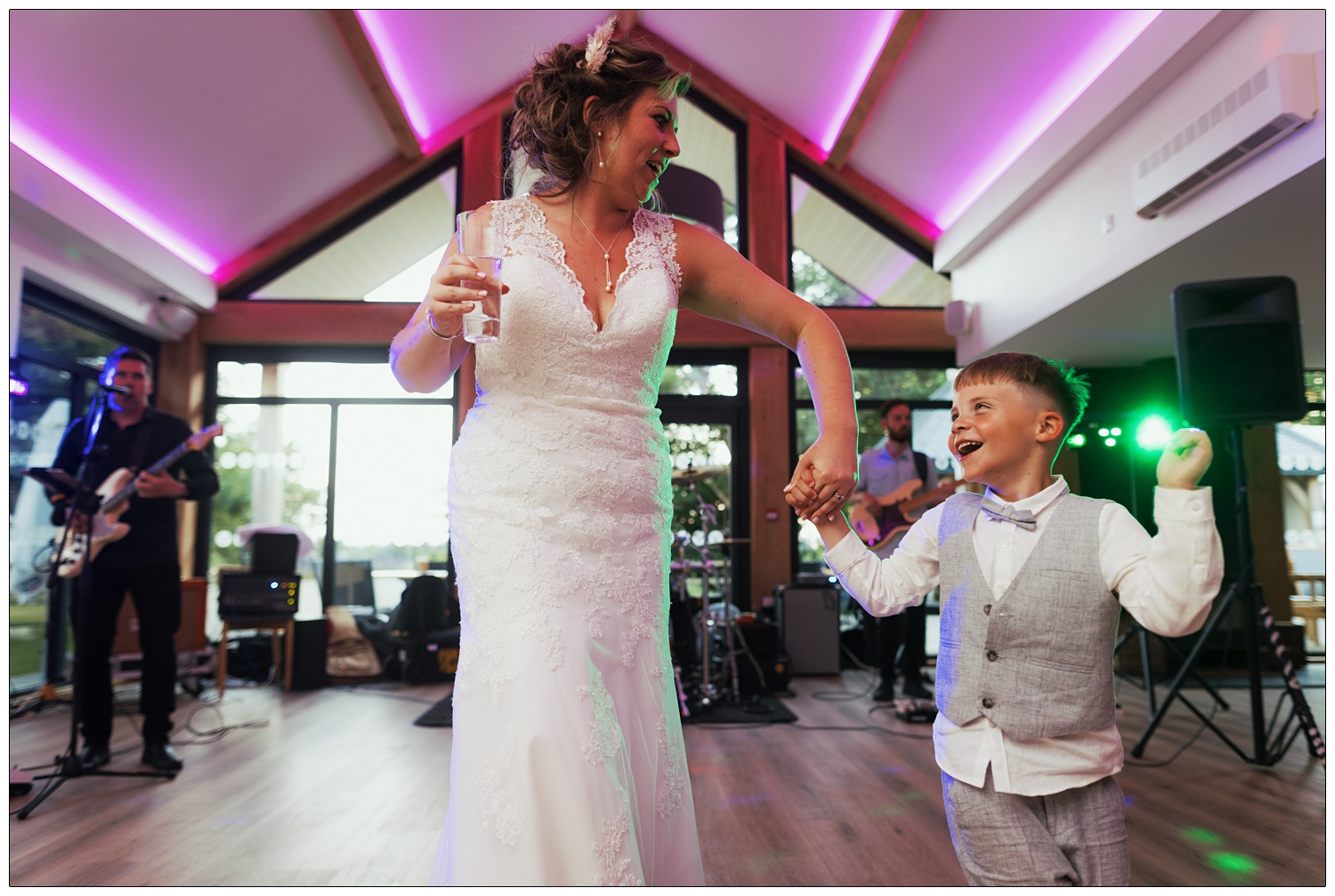 A woman in a lace wedding dress is holding a drink and her nephew's hand. They are dancing at a wedding reception, the lighting is pink and a band are playing behind them.