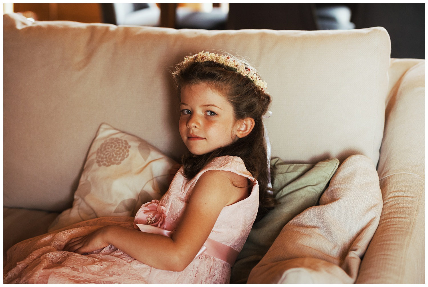 A flower girl sits on a sofa and looks at the camera. She is wearing a pink dress.