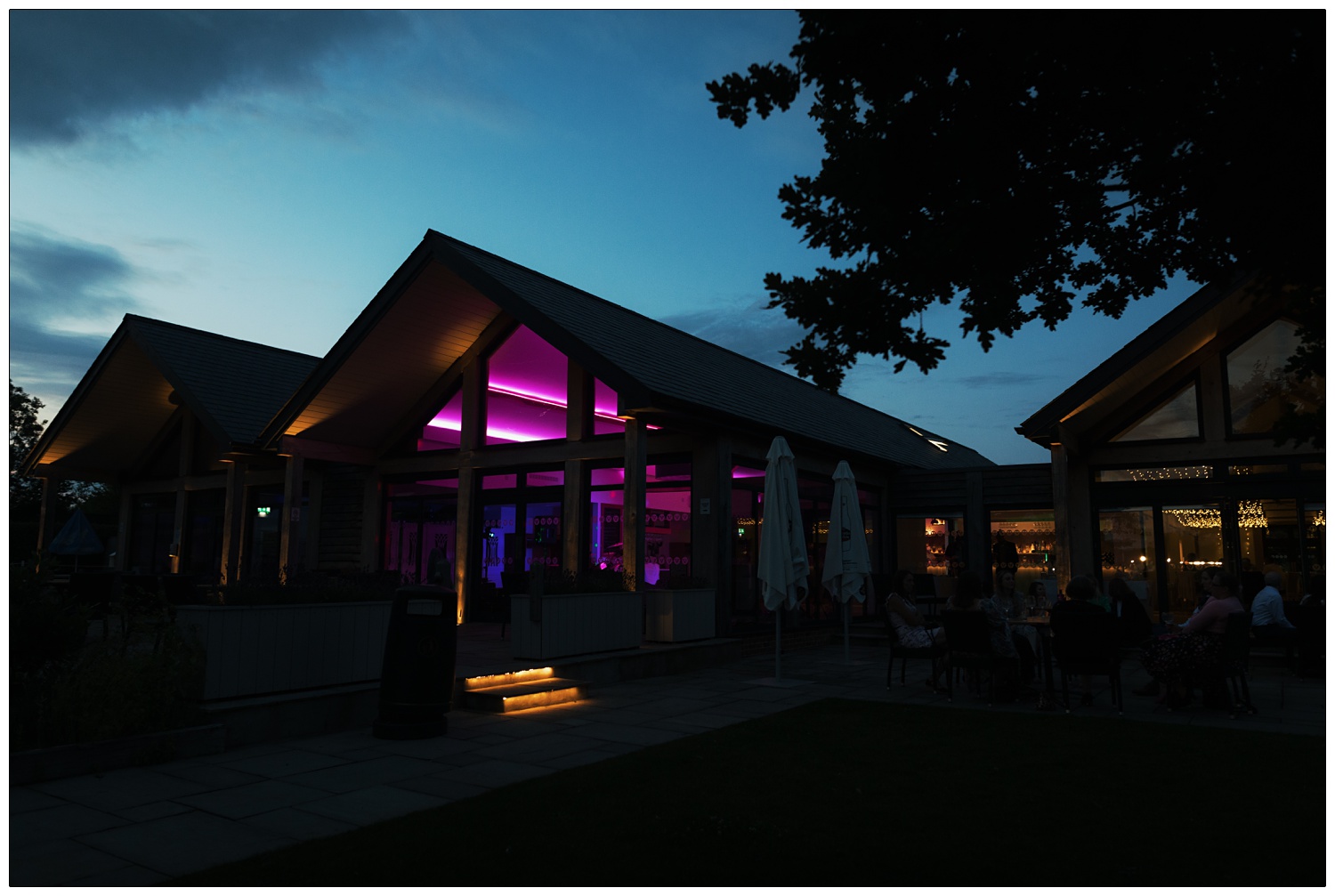 A photograph of Fynn Valley Terrace wedding venue at night. The sky is getting dark, the inside of the venue is lit up magenta.