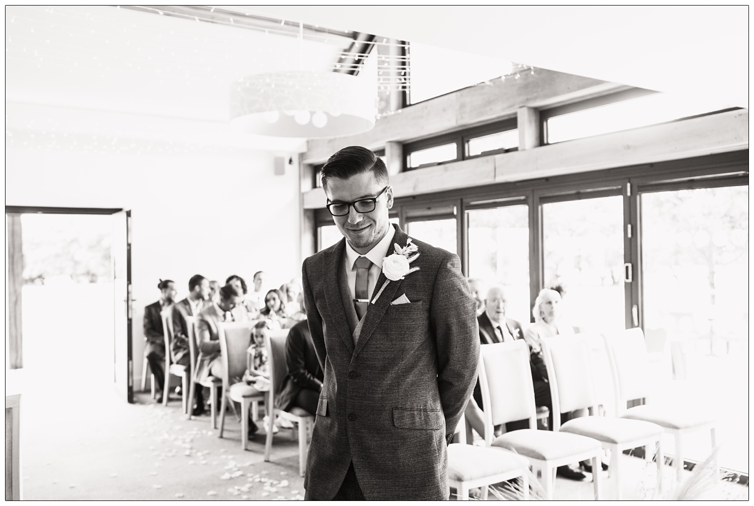 Black and white photograph of the groom waiting for the ceremony to start. The guests are sitting on chairs in the background.
