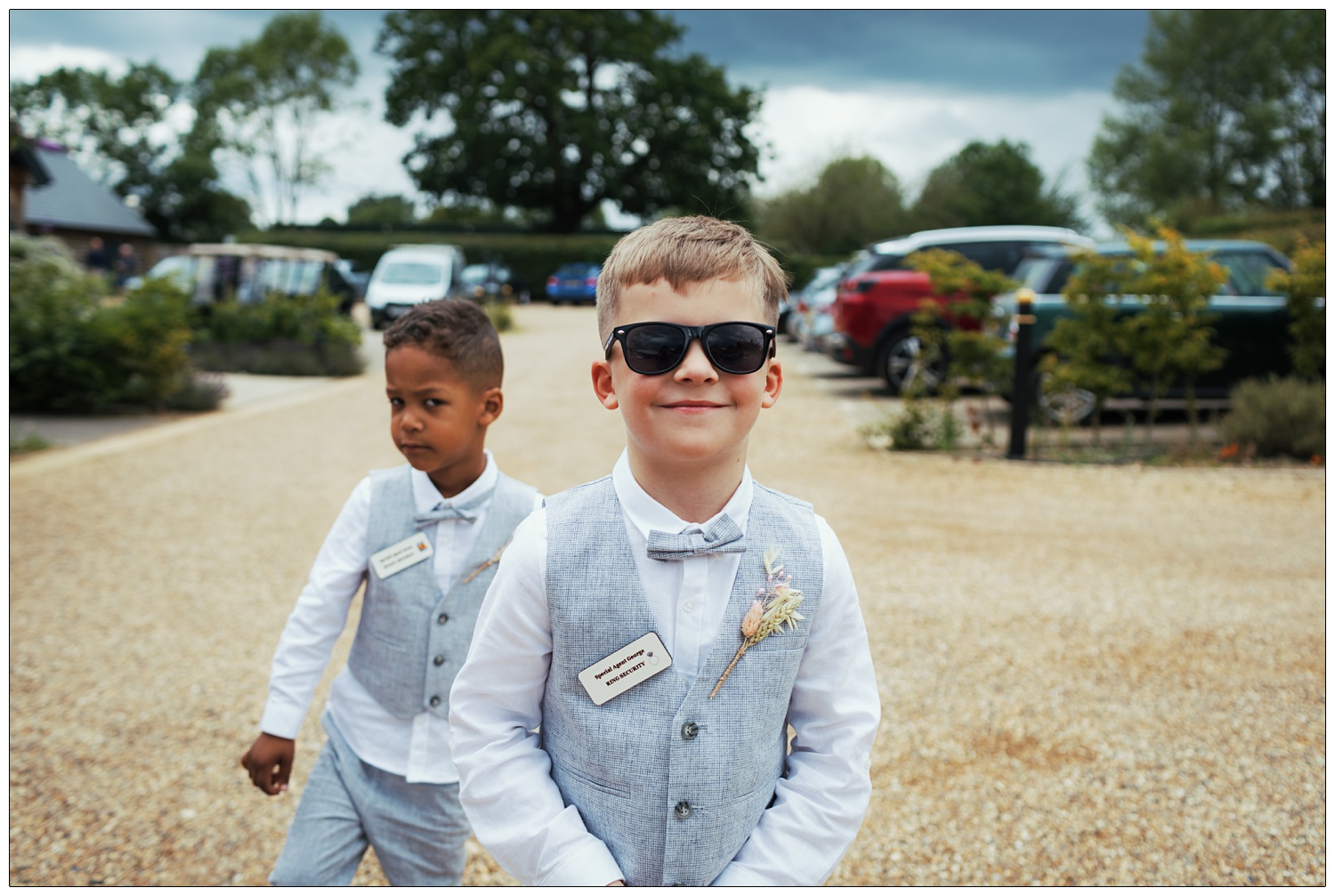 Two young boys in pale blue trousers and waistcoats. They are ring bearers, their badges say "ring security". Boy in foreground is wearing sunglasses.
